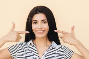 portret van aangenaam ogende brunette vrouw heeft een aantrekkelijke uitstraling, geeft aan met beide voorvingers naar de mond een zachte glimlach te tonen, tanden met beugels, gekleed in vrijetijdskleding, geïsoleerd op een lichte muur foto