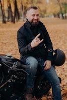 knappe bebaarde mannelijke motorrijder houdt helm vast, maakt hoorngebaar met vingers, voelt cool aan, draagt zwarte jas en spijkerbroek, zit op snelle motor tegen herfstparkachtergrond. zorgeloze racer foto