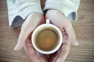 vrouw handen in trui houden een kop van sterk koffie Aan houten tafel. koffie ventilator top visie achtergrond met kopiëren ruimte - wijnoogst stijl foto