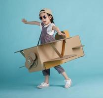 Aziatisch weinig kind meisje spelen met karton speelgoed- vliegtuig handwerk geïsoleerd Aan blauw achtergrond, creatief met familie en dromen van vliegend concept foto