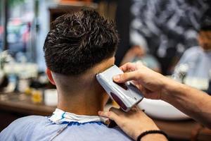 achterzijde visie van jong Mens krijgen een modern kapsel. Mens wezen bijgesneden met professioneel elektrisch tondeuse machine in kapperszaak. mannetje schoonheid behandeling concept. vent krijgen nieuw kapsel in kapper salon foto