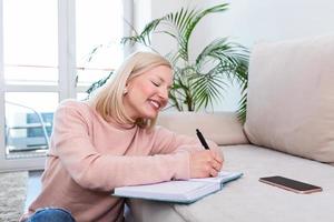 gelukkig albino vrouw zittend Aan de voorzien van vloerbedekking verdieping terwijl aan het studeren haar lessen gebruik makend van een boek met notities. ondernemer of leerling werken of aan het studeren Bij huis en schrijven aantekeningen zittend Aan de verdieping Bij huis foto