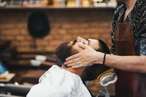 knap Mens met een baard en Gesloten ogen in een zwart snijdend haar- kaap in de kapperszaak. kapper in is aan het doen hem een gezicht massage. detailopname. horizontaal. foto