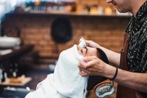 kapper covers de gezicht van een Mens met een heet handdoek. traditioneel ritueel van scheren de baard met heet en verkoudheid comprimeert in een oud stijl kapper winkel. foto