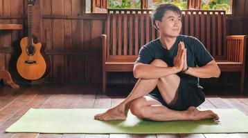 sportief Aziatisch Mens in zwart is aan het doen yoga terwijl oefenen Aan yoga mat Bij huis met houten verdieping.man yoga praktijk houding opleiding gezond leven concept foto