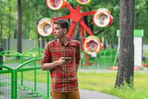 een jong Mens in een plaid overhemd is staand in een park met een telefoon in zijn handen, achter hem is een attractie foto