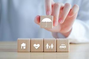 verzekering en bescherming concept. vrouw hand- Holding paraplu icoon met huis, auto, familie en Gezondheid symbool Aan houten blokken voor zekerheid leven concept. foto