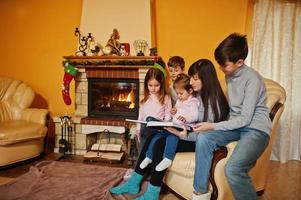 gelukkige jonge grote familie bij een open haard in warme woonkamer op winterdag. moeder met vier kinderen thuis lees boek. foto