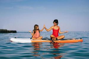 mam en weinig dochter aan het doen yoga Aan de peddelen bord foto