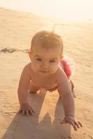 weinig baby meisje is kruipen Aan een zanderig strand in de buurt naar zee in zonsondergang zonlicht. foto