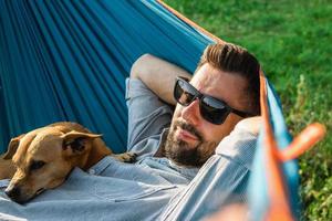 jong knap Europese Mens in zonnebril is resting in hangmat met zijn schattig weinig hond. foto