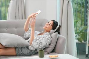 ontspannen vrouw in draadloze hoofdtelefoons luisteren naar muziek- aan het liegen Aan sofa binnen. weekend Bij huis concept foto