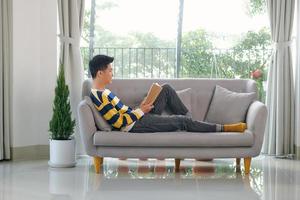 portret van blij jong Mens lezing boek terwijl zittend Aan sofa in zijn leven kamer foto