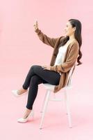 vol lengte portret van prachtig gelukkig vrouw in bedrijf slijtage nemen selfie Aan cel telefoon terwijl zittend Aan stoel foto
