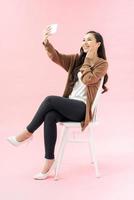 vol lengte portret van prachtig gelukkig vrouw in bedrijf slijtage nemen selfie Aan cel telefoon terwijl zittend Aan stoel foto
