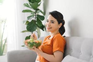 jong vrouw ontspannende Aan de bankstel Bij huis en aan het eten een vers tuin salade, gezond levensstijl en voeding concept foto