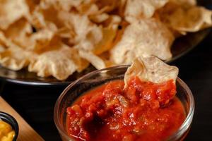 dichtbij omhoog hand- houden spaander dip tomaat salsa met bord met taco, nacho's chips en tomaat duik. pittig nacho's Aan rustiek houten achtergrond foto