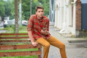 een jong Mens in een plaid overhemd zit Aan een houten traliewerk, houdt een telefoon in zijn handen en types een bericht foto
