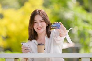 een Aziatisch vrouw zit in een tuin ontspannende met haar mobiel telefoon voor online boodschappen doen en credit kaart betalingen foto