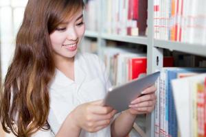 Aziatische mooie vrouwelijke student die tablet in bibliotheek gebruiken foto