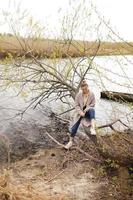 jong mooi vrouw in beige jas is zittend Aan een oud boom in de buurt de rivier- Aan vroeg voorjaar dag tijd foto