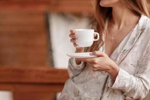 jong vrouw Aan de balkon Holding een kop van koffie of thee in de ochtend. ze in hotel kamer op zoek Bij de natuur in zomer. meisje is gekleed in elegant nachtkleding. kom tot rust tijd foto