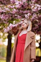 elegant blond in een rood jurk in de voorjaar stad. mooi meisje in rood jurk poseren Aan stad straten. verbijsterend jong vrouw wandelen in de stad foto