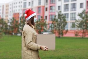 een mooi vrouw in een rood de kerstman claus hoed en medisch beschermend masker houdt een groot doos buitenshuis. nieuw jaar Kerstmis pakket in doos, levering online op te slaan in quarantaine tijd. onderhoud coronavirus. foto