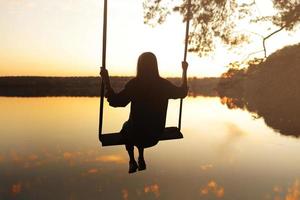 romantische jonge vrouw op een schommel over het meer bij zonsondergang. jong meisje reiziger zittend op de schommel in de prachtige natuur, uitzicht op het meer foto