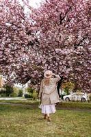 mooi vrouw staat met haar terug in de buurt de sakura bomen. vrouw in hoed, jurk en elegant jas. roze bloemen bloeiend in Oezjhorod, Oekraïne. bloesem in de omgeving van. voorjaar tijd concept foto