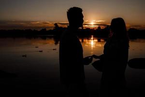 silhouet van liefhebbend paar knuffelen Aan de meer Bij zonsondergang. mooi jong paar in liefde wandelen Aan de kust van de meer Bij zonsondergang in de stralen van helder licht. kopiëren ruimte. selectief focus. foto
