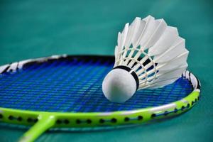 roomwitte badmintonshuttle en racket met neonlichtschaduw op groene vloer in indoor badmintonveld, vage badmintonachtergrond, kopieerruimte. foto