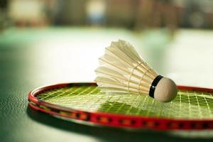 roomwitte badmintonshuttle en racket met neonlichtschaduw op groene vloer in indoor badmintonveld, vage badmintonachtergrond, kopieerruimte. foto