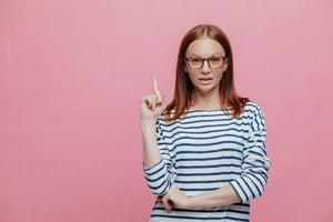 attente mooie vrouw draagt een bril en een gestreepte trui, wijst met de wijsvinger naar boven, staat tegen de roze studiomuur met vrije ruimte voor uw advertentie of promotionele inhoud foto