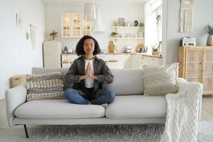 gemengd ras meisje praktijken yoga, meditatie Bij huis zittend in lotus houding Aan bank. gezond levensstijl foto