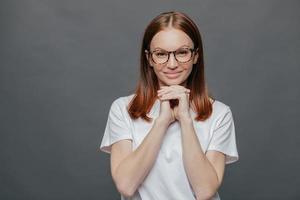 vrolijke aangenaam ogende dame houdt beide handen onder de kin, glimlacht positief naar de camera, draagt een optische bril, casual t-shirt, geniet van vrije tijd in goed gezelschap, geïsoleerd over grijze achtergrond foto