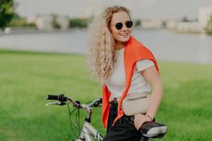 actief levensstijlconcept. positieve jonge vrouw met licht krullend haar, fietst op groen gazon, draagt zonnebril, wit t-shirt en rode trui op schouders, draagt tasje, goed humeur foto