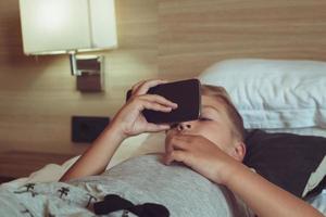 klein jongen aan het kijken tekenfilms Aan slim telefoon terwijl aan het liegen Aan de bed. foto