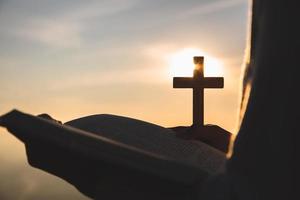 silhouet van jong vrouw handen Holding heilig Bijbel en optillen van christen kruis met licht zonsondergang achtergrond. christen concept foto