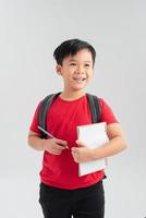 jong Aziatisch school- jongen schrijven in kladblok met rugzak Aan wit achtergrond foto