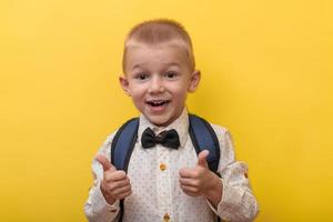 terug naar school. een blond grappig jongen met een rugzak in een licht overhemd Aan een geel achtergrond shows super, klasse en glimlacht. looks Bij de camera. kopiëren ruimte. onderwijs. foto