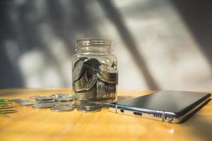 besparing concept een glas van munten en telefoon Aan houten tafel en wazig achtergrond foto