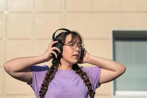 een tiener meisje staat Aan de straat, persen hoofdtelefoons naar haar oren met haar handen en luistert naar muziek- met haar ogen Gesloten. foto