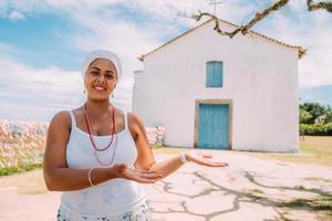 gelukkige braziliaanse vrouw gekleed in traditioneel bahiaans kostuum met iets op de palm van haar hand, kijkt naar de camera, met het historische centrum van porto seguro op de achtergrond foto