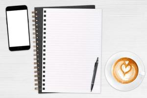 een open blanco notitieboekje, smartphone met pen en een kopje latte koffie op houten tafel. foto