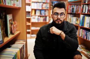 lange slimme arabische studentenman, draag een zwart spijkerjack en een bril, in de bibliotheek met een boek bij de hand. foto