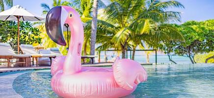 zomer pret in tropisch toevlucht, stoelen, ligstoelen met paraplu onder palm bomen. roze opblaasbaar flamingo zwemmen zwembad water, zomer strand panorama. luxe levensstijl reis, zomer vakantie banier foto