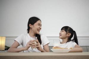 een gezond Aziatisch Thais familie, weinig dochter, en jong moeder drinken vers wit melk in glas en brood vreugde samen Bij een dining tafel in ochtend, welzijn voeding huis ontbijt maaltijd levensstijl. foto