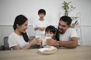 een gezond Aziatisch Thais familie, weinig kinderen, en jong ouders drinken vers wit melk in glas en brood vreugde samen Bij een dining tafel in ochtend, welzijn voeding huis ontbijt maaltijd levensstijl. foto