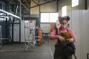een vrouw lasser voorbereidingen treffen voor een werken dag in de metaal verwerken industrie draagt een masker Aan haar gezicht ten gevolge naar de coronavirus pandemisch foto
